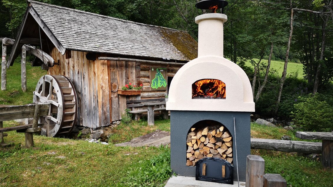 Ein schöner Außenofen aus Stein mit Holzlager steht in einem malerischen Garten. Im Hintergrund sieht man eine rustikale Holzscheune und grüne Bäume. | © Naturpark Sölktäler
