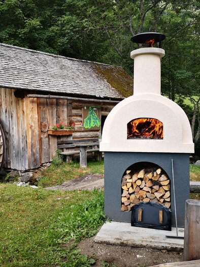 A beautiful stone outdoor oven with a wood storage stands in a picturesque garden. In the background, you can see a rustic wooden barn and green trees. | © Naturpark Sölktäler