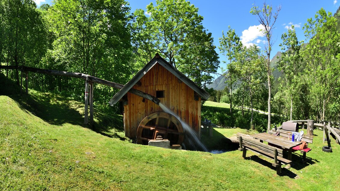 Eine alte Mühle steht in einer grünen Landschaft, umgeben von Bäumen. Im Vordergrund befindet sich eine Holzbank unter dem blauen Himmel. | © Naturpark Sölktäler
