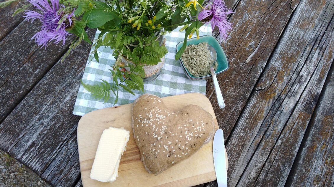 Ein herzförmiges Brot liegt auf einem Holzbrett neben einer Portion Butter. Im Hintergrund steht ein Blumenstrauß und es gibt ein Kästchen mit Kräutern. | © Naturpark Sölktäler