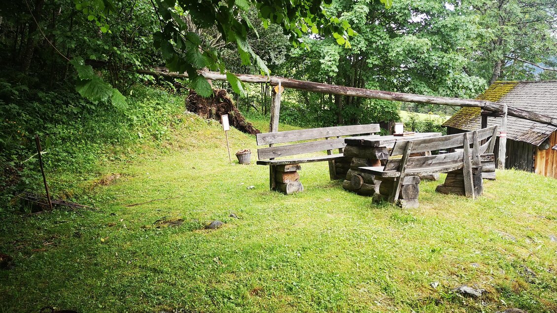 Eine gemütliche Sitzecke aus Holz auf einer grünen Wiese. Im Hintergrund sind Bäume und ein altes Gebäude zu sehen. | © Naturpark Sölktäler