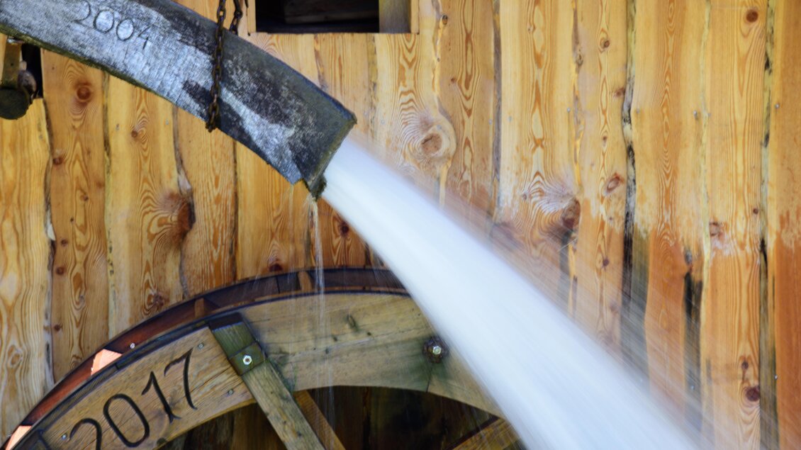 Ein Holzrad mit Wasser, das von einem Auslass fließt. Der Hintergrund besteht aus einer Holzwand mit eingegrabenen Jahreszahlen. | © Naturpark Sölktäler