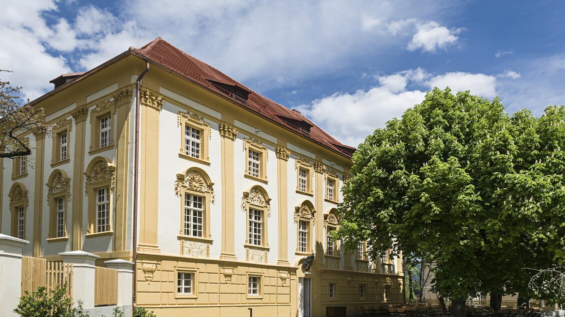 Ein historisches Gebäude mit yellowfarbenen Wänden und kunstvollen Details. Im Vordergrund befindet sich ein großer, grüner Baum und ein klarer Himmel. | © Tauroa GmbH