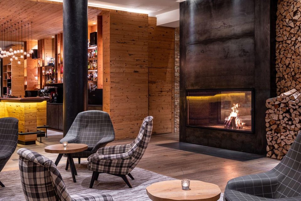Gemütliche Hotellounge mit modernen Sesseln in Karomuster, kleinen Holztischen, einer beleuchteten Bar im Hintergrund und einem großen Kaminfeuer neben gestapeltem Brennholz. | © Fotograf Dragan Dokmanovic