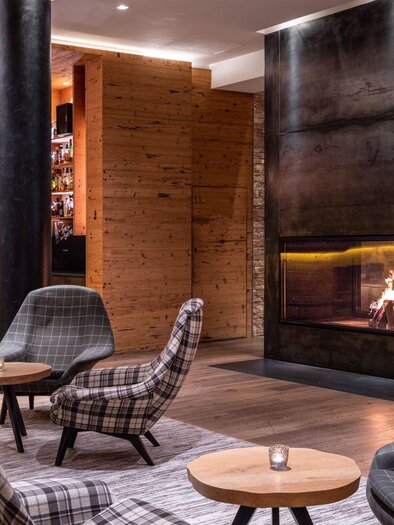 Gemütliche Hotellounge mit modernen Sesseln in Karomuster, kleinen Holztischen, einer beleuchteten Bar im Hintergrund und einem großen Kaminfeuer neben gestapeltem Brennholz. | © Fotograf Dragan Dokmanovic