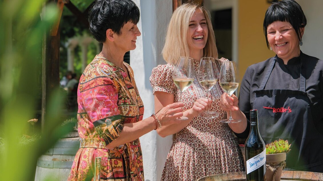 Drei Frauen stoßen mit Gläsern an, während sie draußen stehen. Im Hintergrund ist ein schöner, sonniger Tag zu sehen. | © Weinschloss Thaller