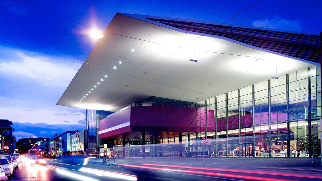 Ein modernes Gebäude mit großen Glasfenstern und futuristischer Architektur. Die Beleuchtung bei Dämmerung schafft eine einladende Atmosphäre. | © MCG - Werner Krug