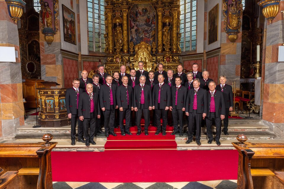 Eine Männerchorgemeinschaft steht in eleganter Kleidung vor einem prunkvollen Altar in einer Kirche. Im Hintergrund sind kunstvoll gestaltete Fenster und goldene Verzierungen zu sehen. | © Carinthia Chor Millstatt