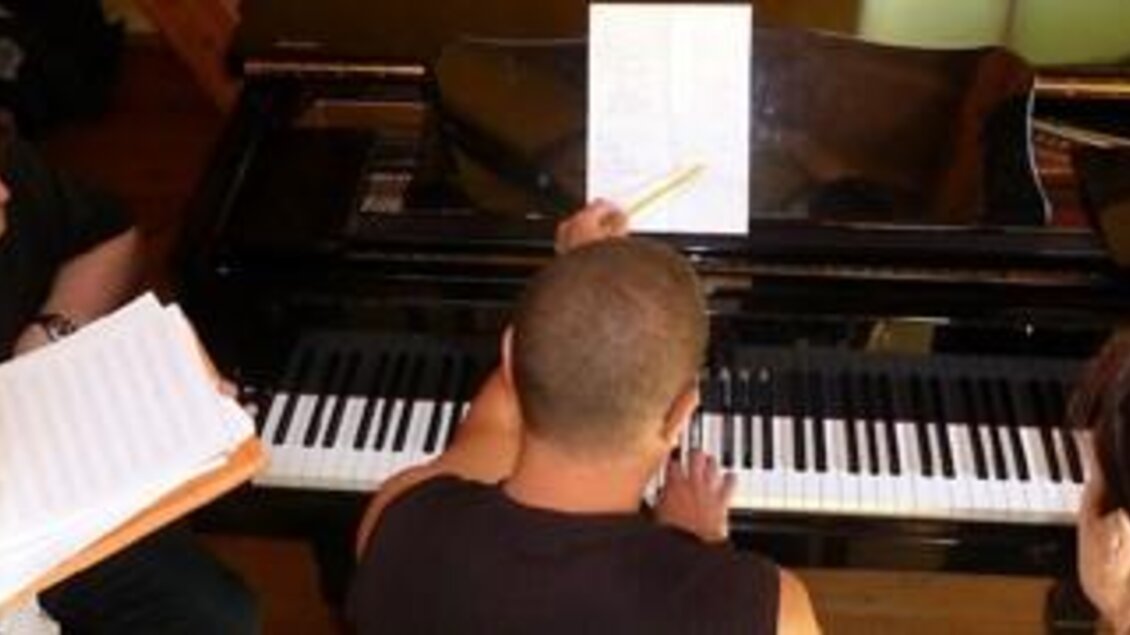 Eine Gruppe von Menschen sitzt um ein Klavier und diskutiert Notizen. Der Fokus liegt auf einem Blatt Papier, das auf der Klaviatur liegt. | © Tom Hejan