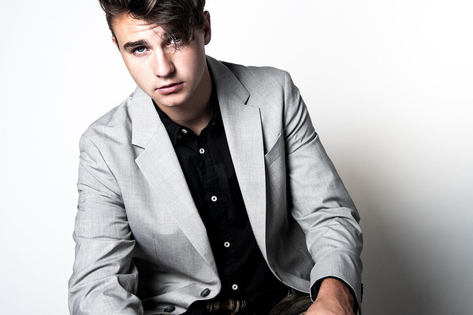 Ein junger Mann sitzt mit einem grauen Blazer und einem schwarzen Hemd. Der Hintergrund ist schlicht und hell, was den Fokus auf ihn lenkt. | © Sasa Felsbach