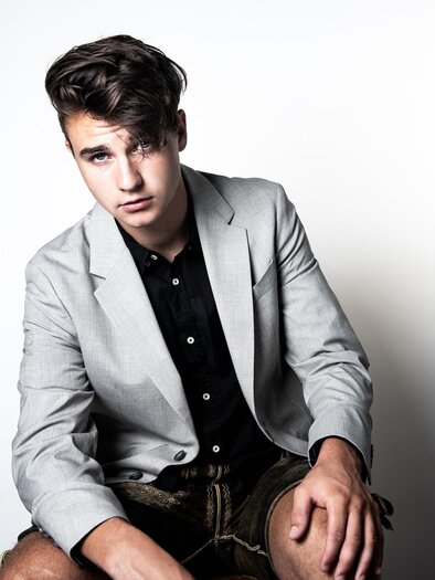 A young man is sitting in a gray blazer and a black shirt. The background is simple and light, which directs the focus on him. | © Sasa Felsbach
