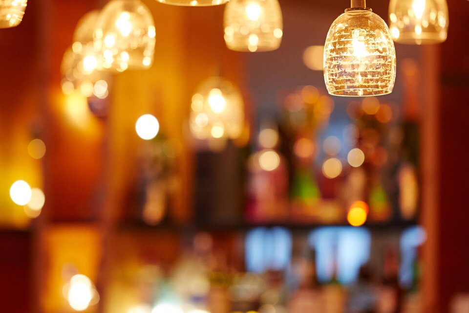 Eine gemütliche Bar mit stilvollen Hängelampen und einer Vielzahl von Getränken im Hintergrund. Die warme Beleuchtung schafft eine einladende Atmosphäre. | © Falkensteiner Hotel Schladming - Fotograf Walter Luttenberger