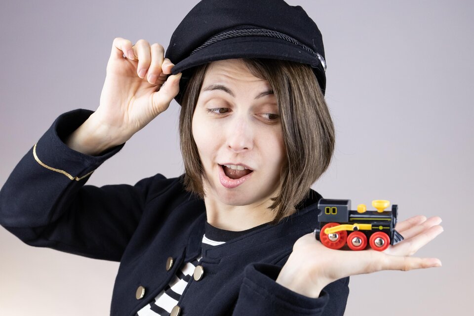 Eine Person mit einem Kapitänshut hält begeistert ein kleines Spielzeugzugmodell in der Hand. Sie trägt ein gestreiftes Shirt und hat eine fröhliche Ausstrahlung. | © Jules Stipsits
