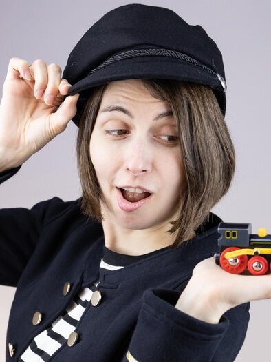 Eine Person mit einem Kapitänshut hält begeistert ein kleines Spielzeugzugmodell in der Hand. Sie trägt ein gestreiftes Shirt und hat eine fröhliche Ausstrahlung. | © Jules Stipsits