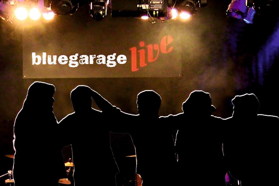 Silhouetten von fünf Personen stehen vor einer Bühne. Im Hintergrund ist ein beleuchtetes Banner mit der Aufschrift "bluegarage live" zu sehen.