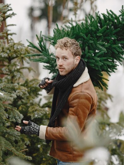 Ein Mann trägt einen künstlichen Weihnachtsbaum und steht zwischen weiteren Bäumen. Die Umgebung wirkt winterlich und festlich. | © freepik