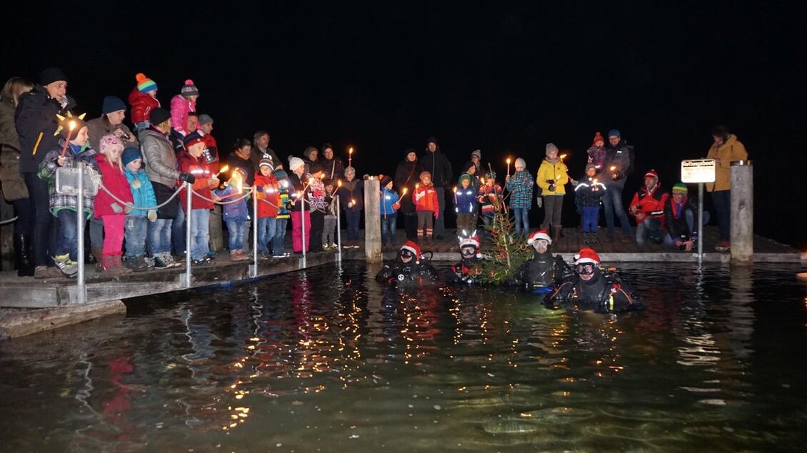 Eine Gruppe von Menschen steht am Ufer und hält Fackeln in der Hand. Im Wasser sind zwei Taucher mit Weihnachtsmützen zu sehen. | © Michael Krainer