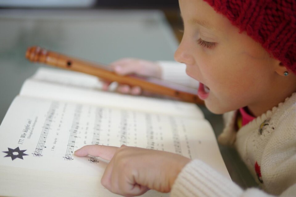 Ein kleines Mädchen spielt Flöte und liest aus einem Musikbuch. Sie trägt eine rote Mütze und zeigt auf die Noten. | © Steirisches Volksliedwerk