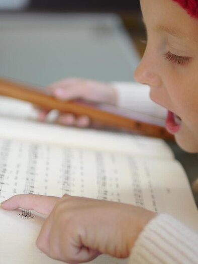 A little girl is playing the flute and reading from a music book. She is wearing a red hat and pointing at the notes. | © Steirisches Volksliedwerk