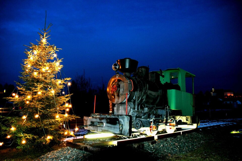 Eine alte Lokomotive steht neben einem geschmückten Weihnachtsbaum unter einem dunklen, blauen Himmel. Die Lichter des Baumes leuchten warm und schaffen eine festliche Atmosphäre. | © Riffel Helmuth