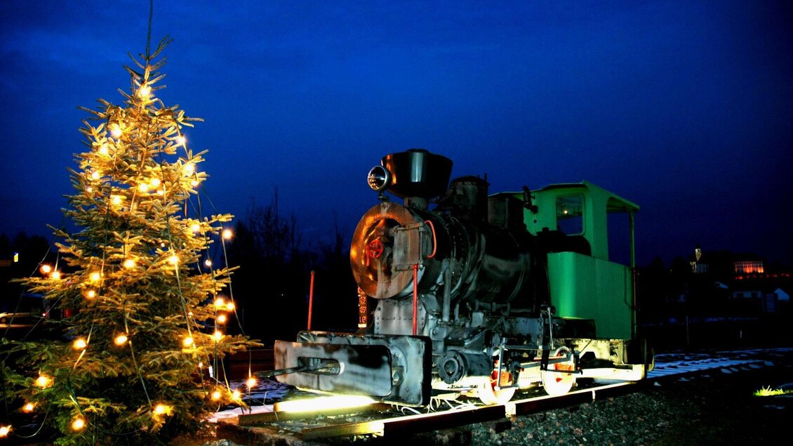 Eine alte Lokomotive steht neben einem geschmückten Weihnachtsbaum unter einem dunklen, blauen Himmel. Die Lichter des Baumes leuchten warm und schaffen eine festliche Atmosphäre. | © Riffel Helmuth