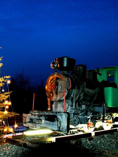 An old locomotive stands next to a decorated Christmas tree under a dark blue sky. The lights of the tree glow warmly, creating a festive atmosphere. | © Riffel Helmuth