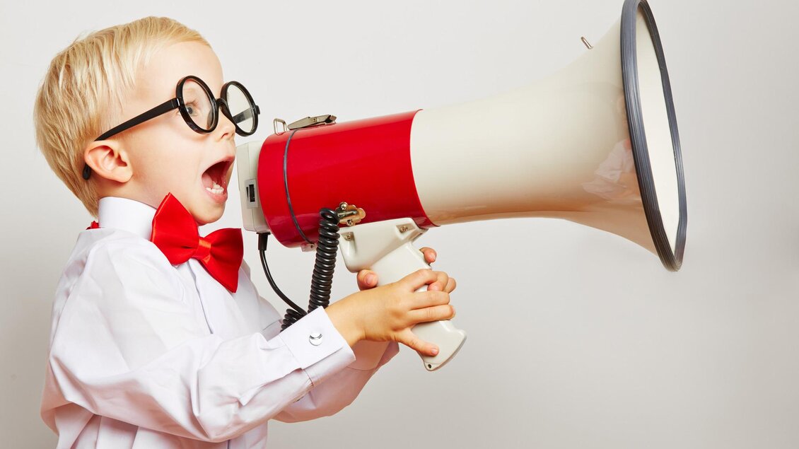 Ein fröhlicher Junge trägt eine weiße Hemd mit einer roten Fliege und großen Brillen. Er schreit in ein Megafon und scheint begeistert zu sein. | © TV Südsteiermark - Robert Kneschke