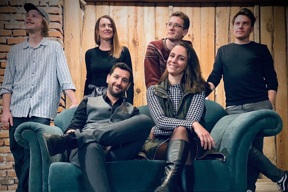Eine Gruppe von sechs Personen sitzt zusammen auf einem Sofa vor einer Holzwand. Alle lächeln und haben eine entspannte Pose. | © Eike Woferlstall