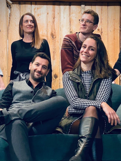 Eine Gruppe von sechs Personen sitzt zusammen auf einem Sofa vor einer Holzwand. Alle lächeln und haben eine entspannte Pose. | © Eike Woferlstall