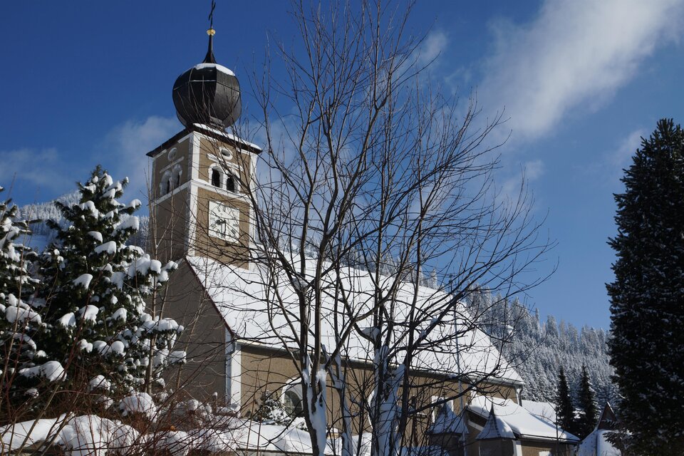 Eine verschneite Kirche mit einem schönen Turm und klar blauem Himmel. Umgeben von schneebedeckten Bäumen und Bergen. | © Alois Leitner