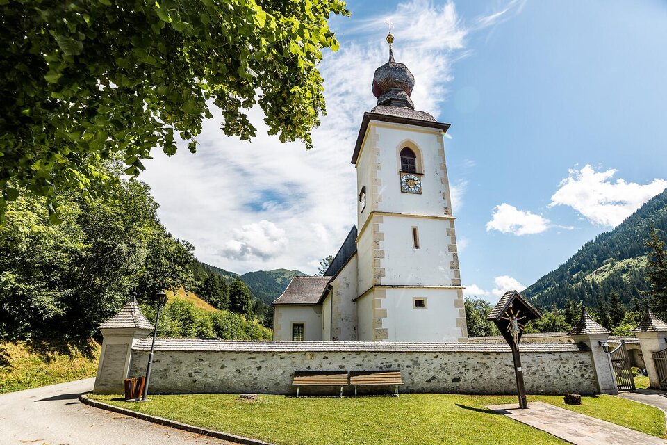 Eine malerische Kirche mit einem hohen Turm umgeben von grünen Bergen. Der Himmel ist klar und die Landschaft ist friedlich.