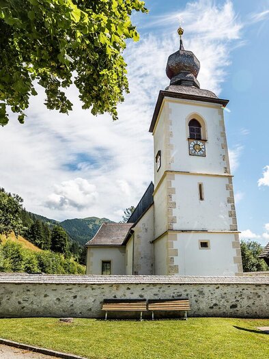 Eine malerische Kirche mit einem hohen Turm umgeben von grünen Bergen. Der Himmel ist klar und die Landschaft ist friedlich.