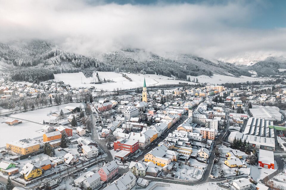 Eine malerische Stadt im Schnee mit bunten Häusern und einem Kirchturm. Die umgebenden Berge sind leicht bewölkt.
