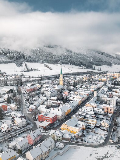Eine malerische Stadt im Schnee mit bunten Häusern und einem Kirchturm. Die umgebenden Berge sind leicht bewölkt.