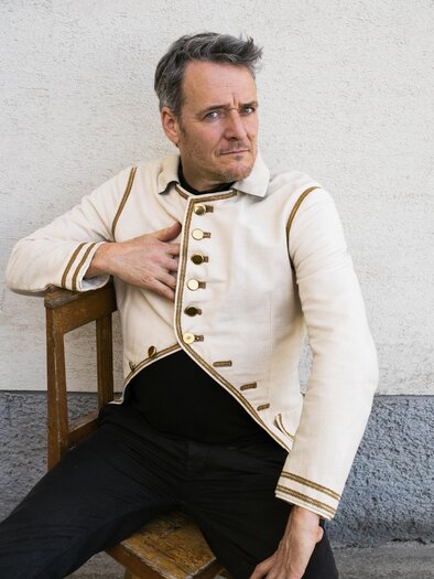 A man is sitting on a chair and wearing a striking, bright jacket with golden buttons. He is looking directly at the camera with a confident expression. | ©  Paul Vincenth Schütz