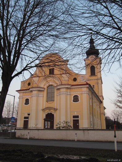 A yellow church with a striking facade and a tall tower. Surrounded by trees and a clear sky.