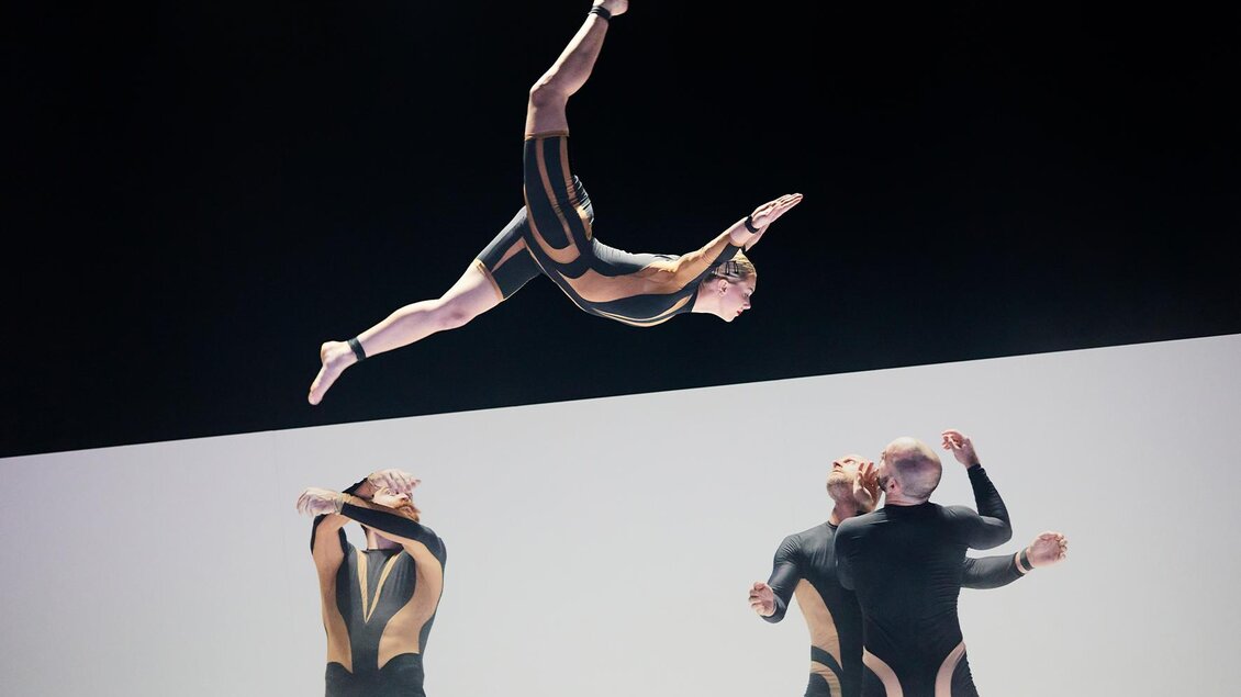 Eine beeindruckende Akrobatik-Darbietung mit einem Künstler, der durch die Luft springt. Im Hintergrund sind drei weitere Akrobaten in action. | © (c)_Andy_Phillipson