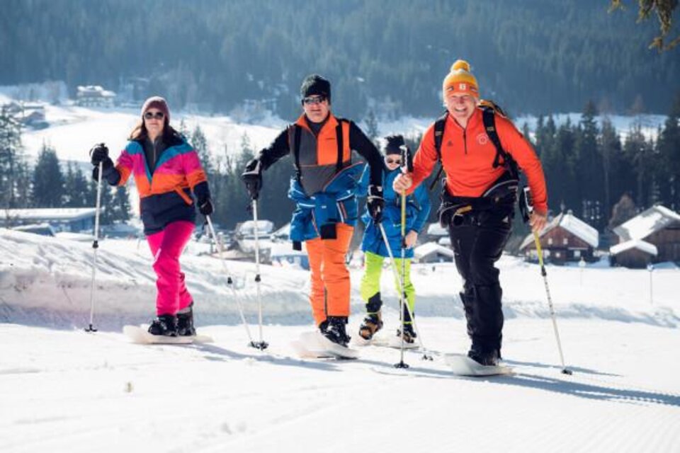 Eine Gruppe von vier Personen macht Schneeschuhwandern in einer verschneiten Landschaft. Im Hintergrund sind Häuser und Tannenbäume sichtbar. | © OCT Outdoor Consulting Group