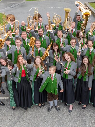 Eine große Musikgruppe steht im Freien und hält ihre Instrumente in die Luft. Die Mitglieder tragen traditionelle Kleidung mit grünen Elementen. | © TMV St. Magdalena a.L.
