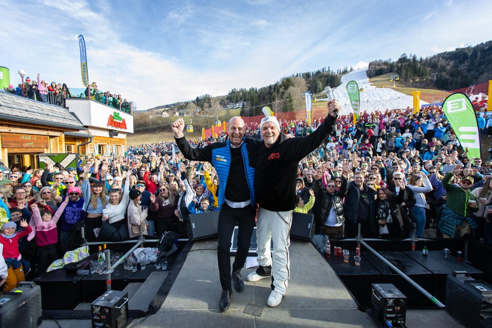 Eine große Menschenmenge feiert im Freien, während zwei Männer auf einer Bühne stehen und die Arme heben. Im Hintergrund sind Berge und ein Skigebiet sichtbar. | © Hauser Kaibling