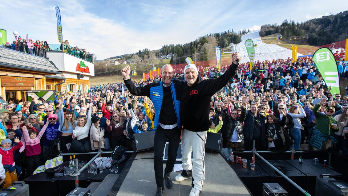 Eine große Menschenmenge feiert im Freien, während zwei Männer auf einer Bühne stehen und die Arme heben. Im Hintergrund sind Berge und ein Skigebiet sichtbar. | © Hauser Kaibling