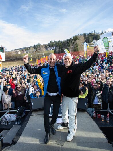 Eine große Menschenmenge feiert im Freien, während zwei Männer auf einer Bühne stehen und die Arme heben. Im Hintergrund sind Berge und ein Skigebiet sichtbar. | © Hauser Kaibling