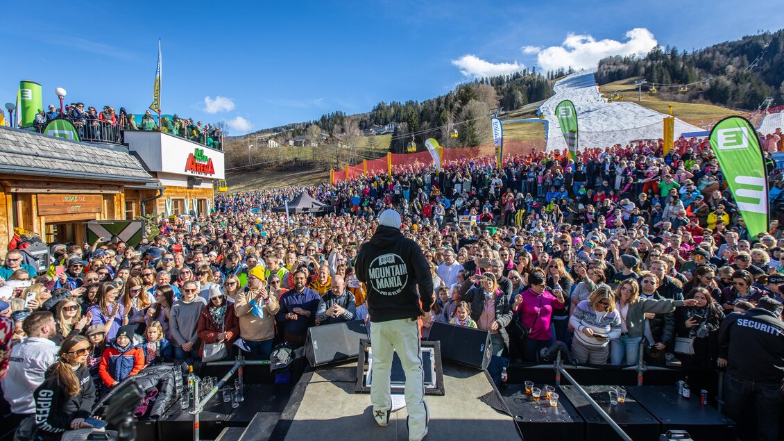 Eine große Menschenmenge versammelt sich vor einer Bühne in den Bergen. Die Veranstaltung findet bei schönem Wetter und klarer Sicht statt.