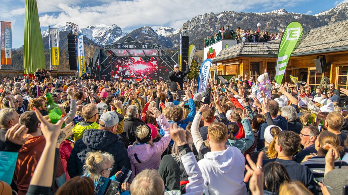 Eine lebhafte Menschenmenge feiert bei einem Konzert in den Bergen. Die Atmosphäre ist festlich, mit bunten Fahnen und einer beeindruckenden Bergkulisse.