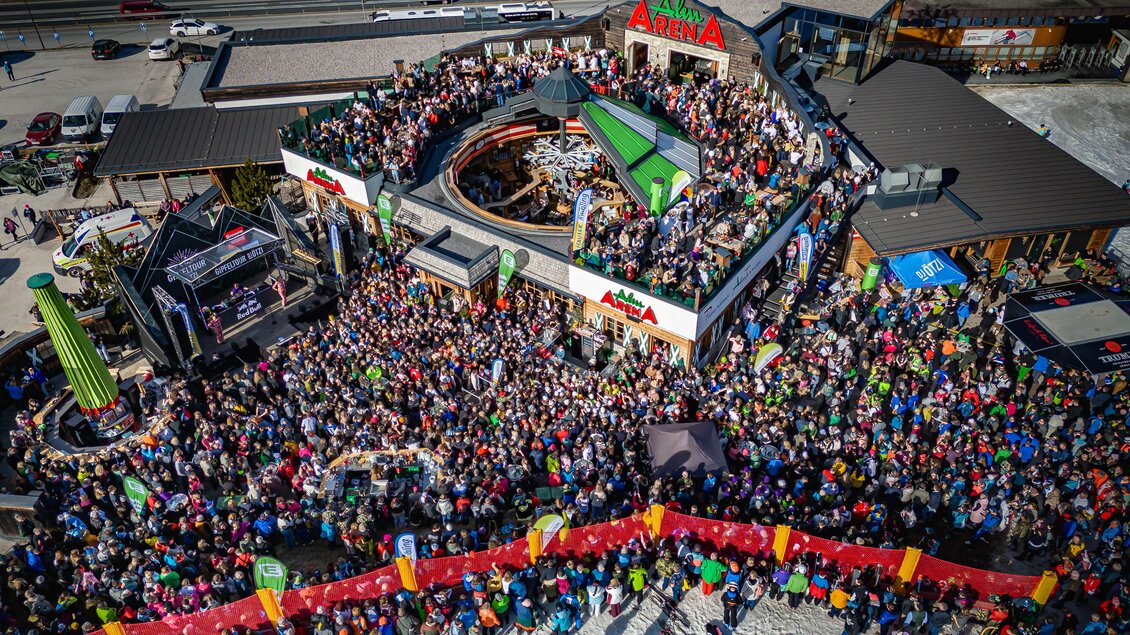 Eine große Menschenmenge versammelt sich vor einem geschäftigen Veranstaltungsort. Bunte Banner und eine Bühne sind sichtbar, was auf eine besondere Veranstaltung hinweist.