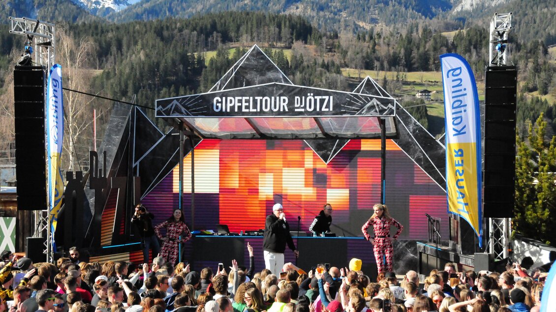 Eine lebhafte Veranstaltung im Freien mit einer großen Menschenmenge vor einer Bühnenaufführung. Im Hintergrund sind beeindruckende Berge und eine klare, sonnige Atmosphäre zu sehen.