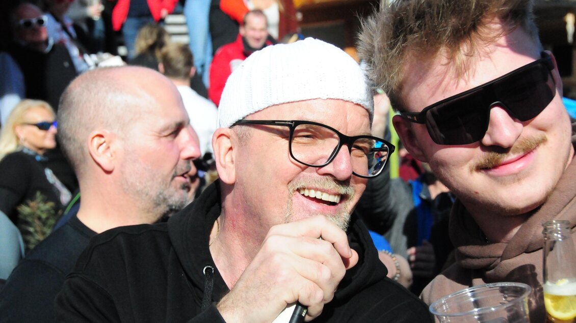 Eine fröhliche Menschenmenge bei einer Veranstaltung im Freien. Zwei Männer lächeln und halten Getränke in der Hand.