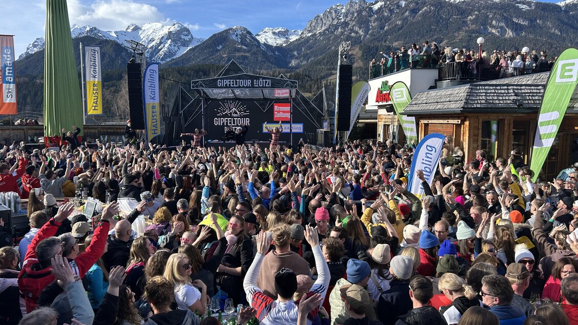 Eine lebhafte Menschenmenge feiert im Freien, umgeben von Bergen und einem klaren blauen Himmel. Im Hintergrund ist eine Bühne mit Bannern und einem musikalischen Event zu sehen.