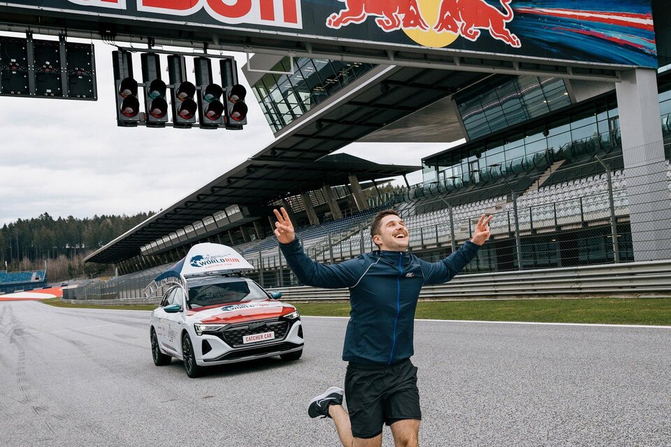 Ein Mann läuft fröhlich auf einer Rennstrecke unter einem Red Bull-Schild. Im Hintergrund ist ein Auto zu sehen, das sich auf der Strecke befindet. | ©  Lucas Pripfl Red Bull Ring