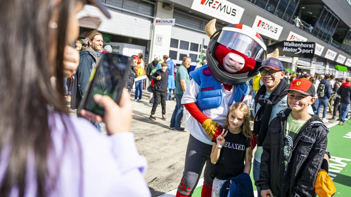 Eine Gruppe von Menschen posiert für ein Foto mit einem maskierten Charakter in einem Rennanzug. Im Hintergrund sind Zuschauer und ein Motorsportgelände zu sehen. | © Red Bull Ring